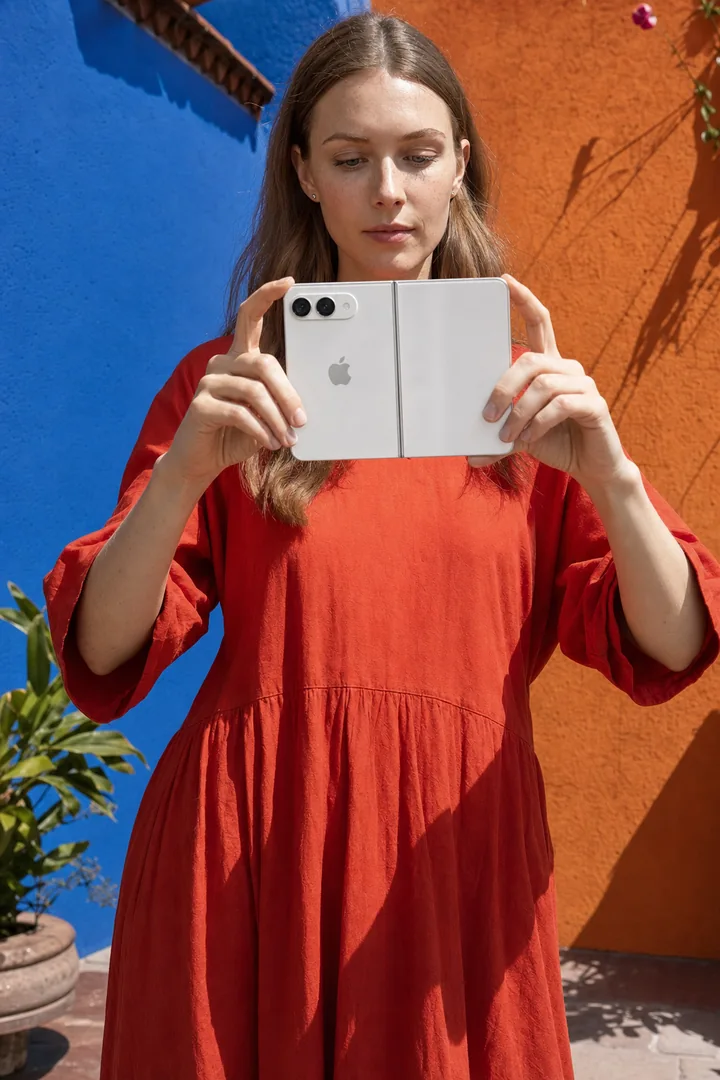 Retrato con teléfono plegable junto a una pared naranja; imagen generada con IA.
